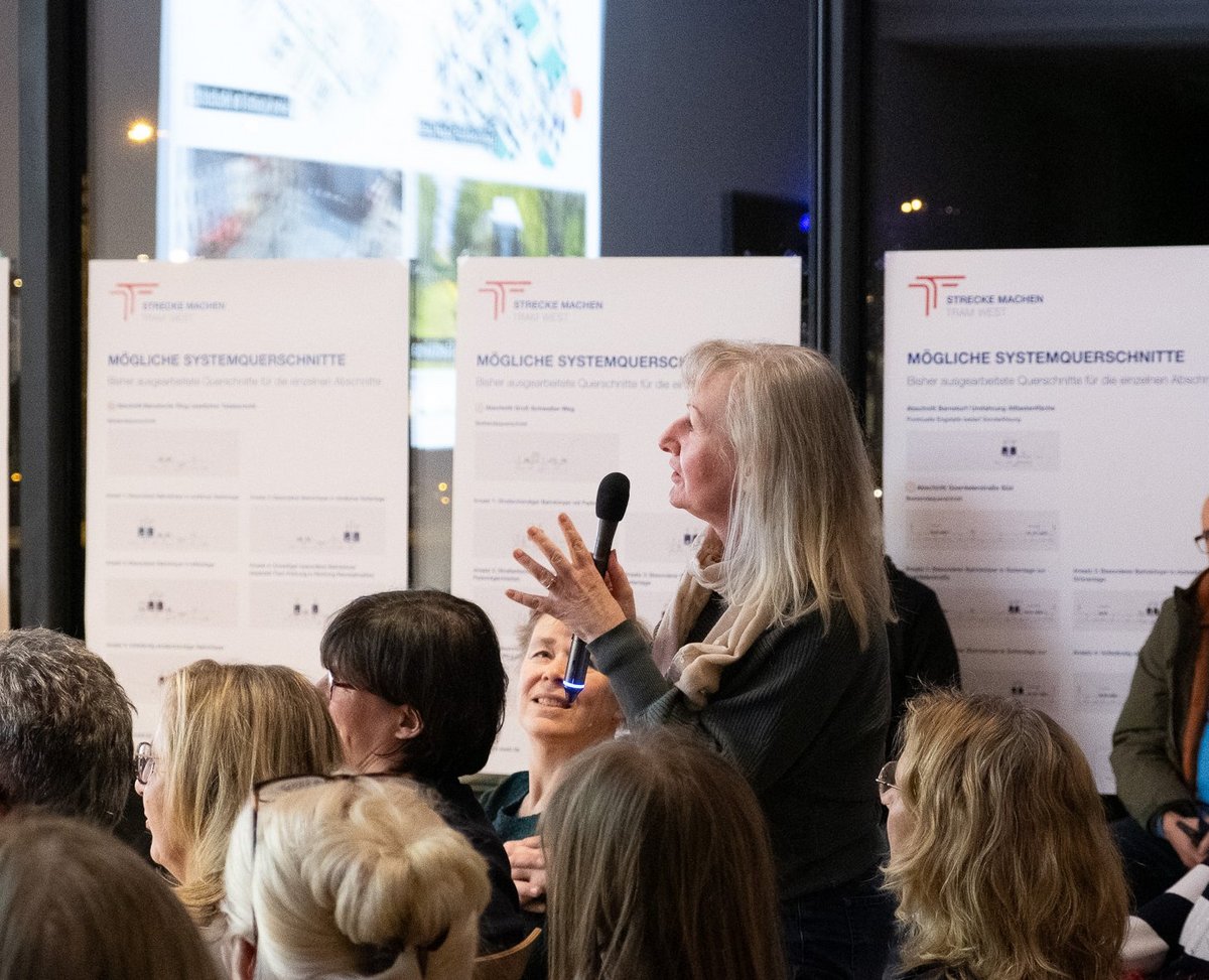 Eine Frau hält ein Mikrofon in der Hand und richtet eine Frage an die Podiumsteilnehmer. Sie steht mitten im Publikum.