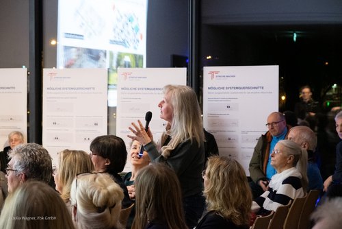 Eine Frau hält ein Mikrofon in der Hand und richtet eine Frage an die Podiumsteilnehmer. Sie steht mitten im Publikum.